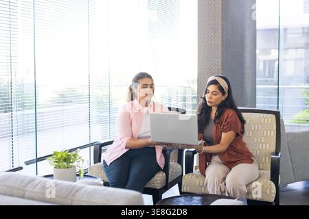 Diverse female colleagues in workwear in office lounge on cushioned chairs reviewing silver laptop Stock Photo