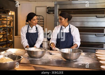Two female bakers kneading dough, talking in chef jackets at floury prep table with bowls, racks Stock Photo