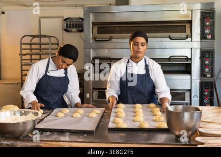 Diverse female bakers in jackets and aprons preparing rolls on stainless steel worktable with ovens Stock Photo