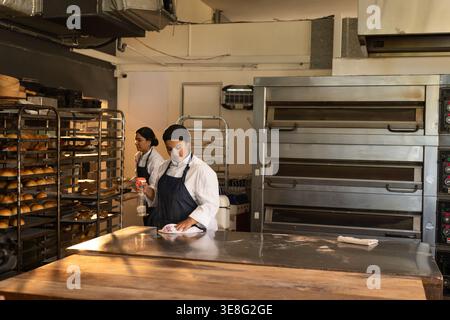 Female bakers cleaning prep table in chef coats using sprayer and organizing rack trays in bakery Stock Photo