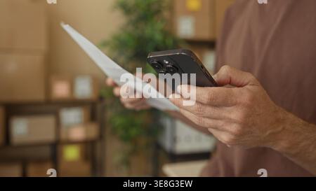 Man holding smartphone and paper in a building, hands visible and stacked boxes nearby; small business focus. Stock Photo