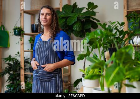 Non-binary shop worker arranging pots and watering houseplants on work counter in striped apron Stock Photo
