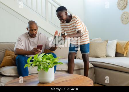 Father and son sitting on sofa, youth holding tablet gesturing at red phone, father checking help Stock Photo