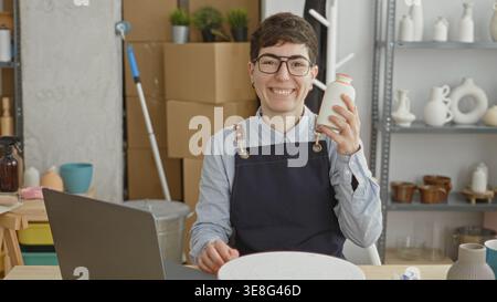 Woman holding ceramic vase on a turntable in a studio while wearing an apron and working on a laptop; craft business creativity. Stock Photo