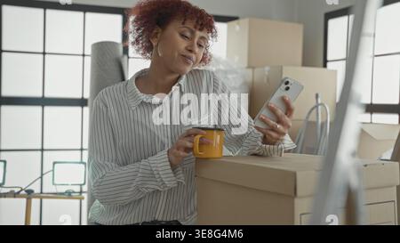 Woman in striped shirt holds smartphone in right hand and yellow mug in left while leaning on moving boxes in building looking at screen; moving day c Stock Photo