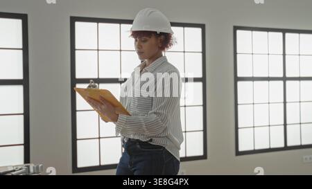 Woman architect in hardhat holds yellow clipboard and inspects an empty house room in building while smiling and checking plans; professional confiden Stock Photo