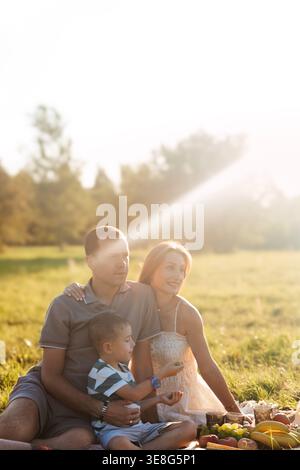 Family gathers on a green field for a picnic, sharing food and smiles while soaking in warm afternoon sunshine, creating joyful memories together. Stock Photo