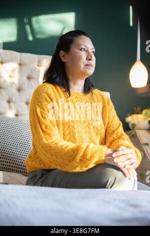 Sitting mature woman gazing right on bed in bedroom, wearing mustard sweater, pendant lamp glowing Stock Photo