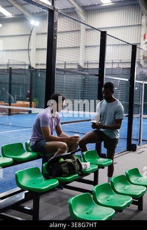 Sitting and standing two players in athletic shirts chatting at court, green seats, duffel, phone Stock Photo