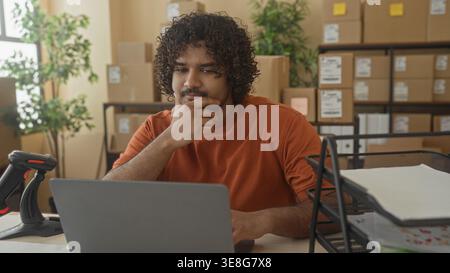 Man in orange shirt with laptop and cardboard box, hand on cheek while sorting paperwork in building; tired small business. Stock Photo