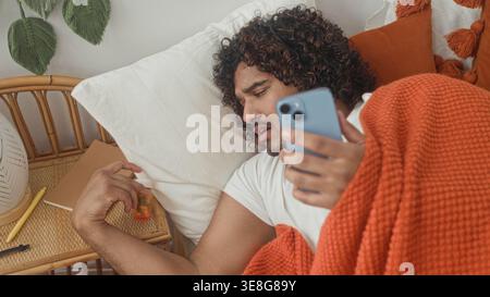 Man in bed holding smartphone, hand on neck and reaching for pill bottle on bedside table, covered by orange blanket, studio; sick. Stock Photo