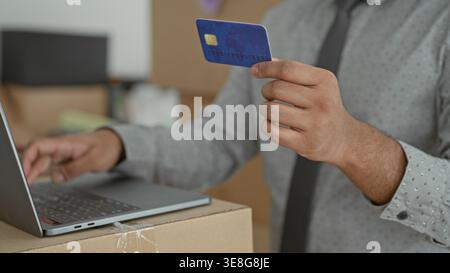 Young indian man holds creditcard in one hand and types on laptop with the other amid moving boxes in a building; convenience. Stock Photo
