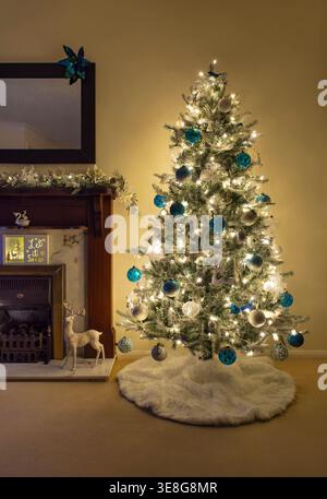 Illuminated Christmas tree at night with blue and white baubles beside fireplace and festive decorations in cosy UK living room interior Stock Photo