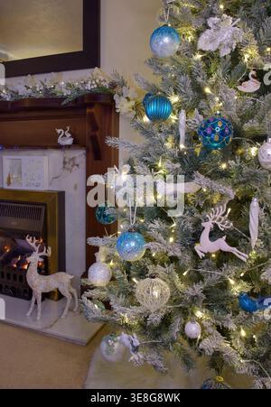 Decorated Christmas tree with blue and white baubles and warm lights beside fireplace in cosy living room, festive seasonal home interior Stock Photo