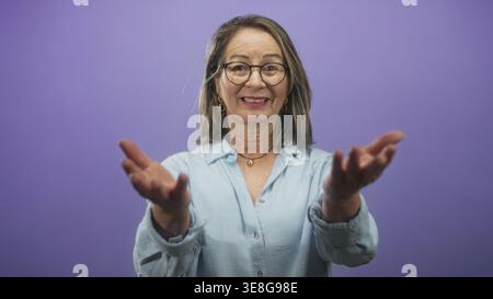 Hispanic woman with grey hair beckons with both hands toward camera in a studio wearing glasses and a light blue shirt; friendly invitation. Stock Photo