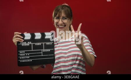 Woman holding clapperboard and showing peace sign in red studio, wearing striped shirt and glasses; joy celebration fun. Stock Photo