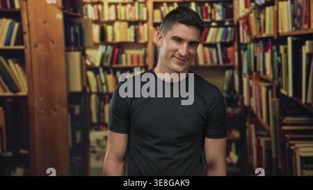 Young man smiling and slightly leaning by library shelves filled with books in a building; calm curiosity. Stock Photo