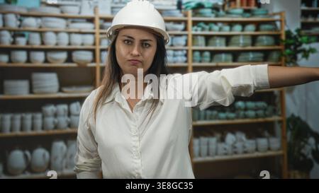 Woman wearing white hard hat with arms crossed and an outstretched arm in home decor shop among ceramic shelves; frustration. Stock Photo