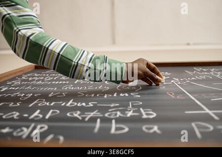 Black Teacher Writing Economics Formula on Chalkboard in Classroom Stock Photo