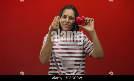 Young hispanic woman holds red heart near face and beckons with hand in studio; pleading love appeal. Stock Photo