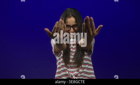 Woman with glasses smiles and beckons with both palms forward wearing a striped tied shirt in a deep blue studio; playful invitation. Stock Photo