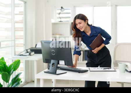 Adult Asian woman stands at desk looking at computer while holding clipboard under her arm. She smiles typing on keyboard in a bright modern office. Stock Photo
