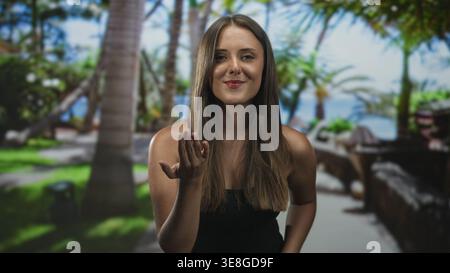 Young hispanic woman with long brunette hair extends palm, beckons with open hand wearing black strapless top in studio; welcoming warmth. Stock Photo