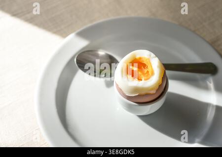 Boiled egg with soft yolk in a stand and a spoon on a white plate on a sunny day, protein snack for breakfast, lunch or dinner, copy space, selected f Stock Photo