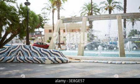 Soft blurred city plaza with fountain, palm trees and mosaic bench in shallow bokeh background; background backdrop copyspace calm. Stock Photo