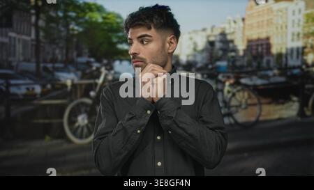 Young hispanic man stands by canal street holding clasped hands under chin with anxious expression and tense posture; fear. Stock Photo