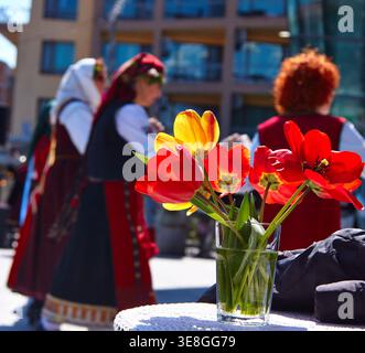 Bulgarian women dressed in traditional costumes gather in an urban area, with a glass vase of colorful tulips prominently displayed in the foreground Stock Photo