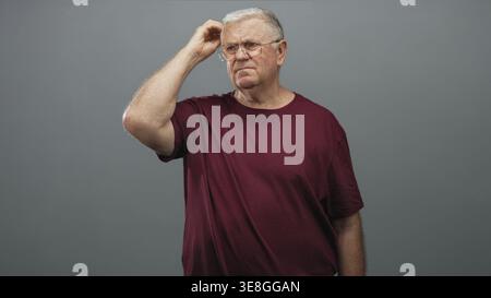 Senior man in burgundy t shirt scratching head with right hand while wearing glasses in studio grey wall setting; confusion reflection pondering. Stock Photo