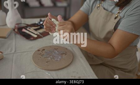 Woman shaping clay with hands at studio table, wearing apron and using sculpting tools and a wooden board; craft skill calm. Stock Photo