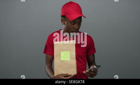 Young man holding smartphone and paper bag with smiley note while looking down in gray studio; curiosity. Stock Photo
