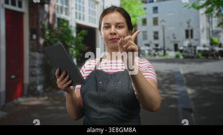 Woman in apron taps tablet on street while checking orders and pointing at tablet screen; focus efficiency. Stock Photo