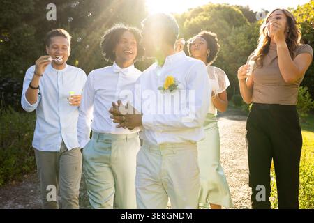 Diverse group of five friends walking along gravel park path, blowing soap bubbles, showing bow tie Stock Photo
