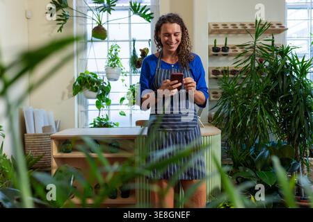 Non-binary African American wearing striped apron at wood counter in plant shop, checking phone Stock Photo