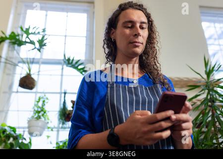 Non-binary plant shop worker wearing striped apron using phone, looking down near kokedama plants Stock Photo