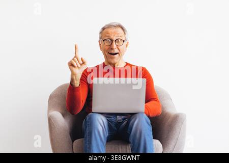 Senior Man Sitting in a Chair Using a Laptop and Expressing Excitement in a Bright Room Stock Photo