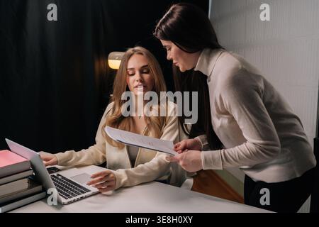 Two businesswomen consult over financial reports and laptop, discussing strategy, sharing ideas and analyzing data during focused office interview and Stock Photo