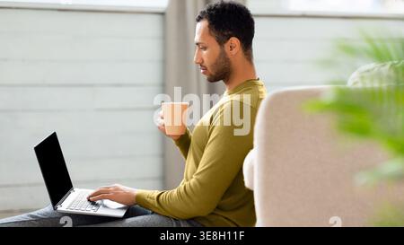 Man Working on Laptop While Sitting on the Floor and Holding a Cup in a Light Room Stock Photo