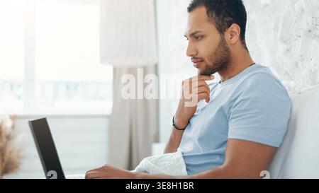 Young Man Working on Computer in Bedroom While Sitting on Bed During Morning Hours Stock Photo