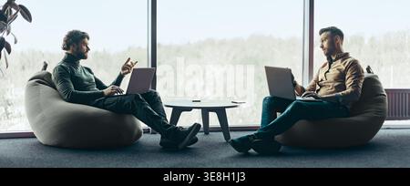 Two Men Sitting in Bean Bag Chairs Talking and Working on Laptops in an Office Space Stock Photo