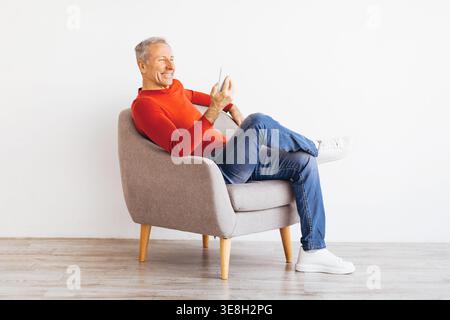 Older Man Sits on Chair and Smiles While Using Smartphone in Indoor Setting Stock Photo