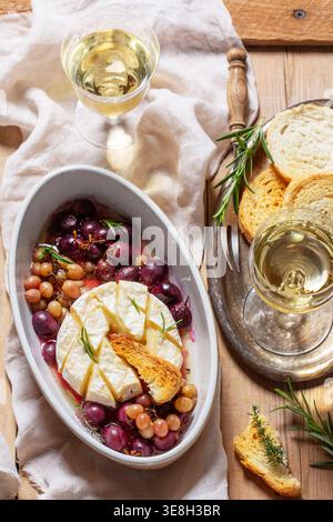 Baked Camembert cheese with grapes, thyme, and rosemary, served with white wine and a baguette. Stock Photo