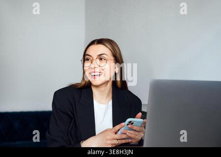 Attractive Business Woman Using Smartphone at Office Desk With Laptop. Corporate Workplace and Communication Concept. Female Professional. Stock Photo