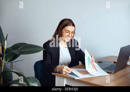 Business Woman Analyzing Charts and Documents at Office Desk With Laptop. Corporate Workplace and Data Analysis Concept. Female Professional. Stock Photo