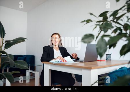 Woman sitting at office desk with laptop and documents. Attractive businesswoman smiling and working in modern corporate workplace. Female professiona Stock Photo