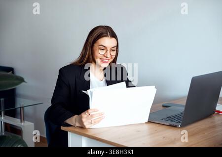 Beautiful Business Woman Holding Documents and Working at Office Desk With Laptop. Corporate Workplace and Paperwork Concept. Stock Photo
