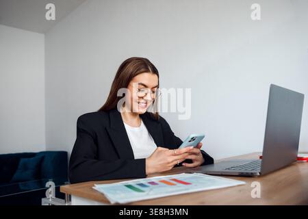 Woman using smartphone at office desk with laptop and documents. Attractive businesswoman working in corporate environment. Female professional multit Stock Photo